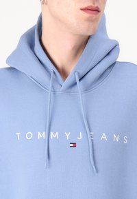 Lichtblauwe hoodie van zachte stof met een trekkoordcapuchon en witte tekst "TOMMYJEANS" met een klein kleurbloklogo.