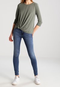 Olivgrünes, langärmliges Oberteil mit einem geknoteten Frontdetail, kombiniert mit eng anliegenden blauen Jeans und weißen Sneakers. Weicher Stoff mit lockerer Passform.