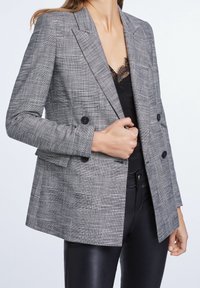 Blazer grigio a check con design doppiopetto, rever a punto, e due bottoni frontali, abbinato a un top in pizzo nero sottostante.