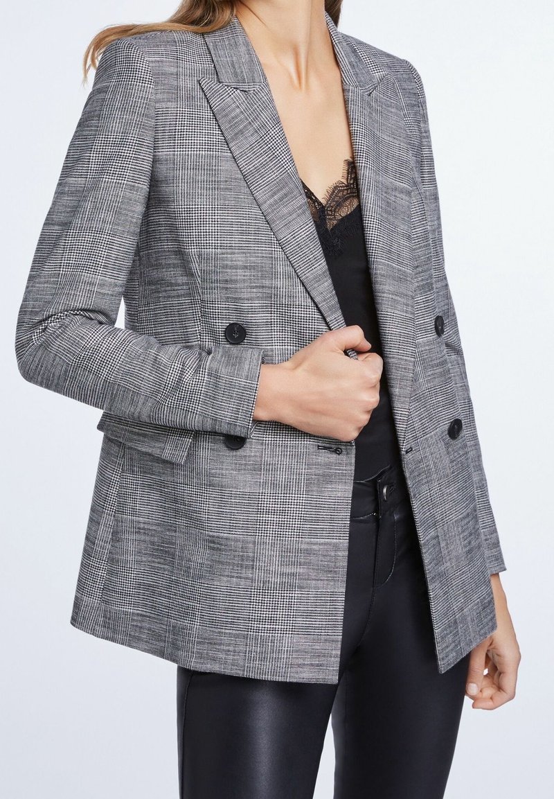Blazer grigio a check con design doppiopetto, rever a punto, e due bottoni frontali, abbinato a un top in pizzo nero sottostante.