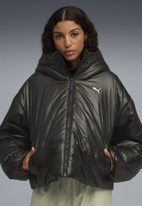 Zwarte pufferjack met een glanzende afwerking, oversized capuchon en ritssluiting. Voorzien van een klein wit logo op de linkerkant van de borst en elastische manchetten.