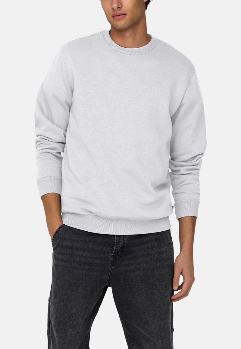 Homme portant un sweat-shirt gris clair uni à col rond et un jean foncé, les mains partiellement dans les poches, sur fond blanc.