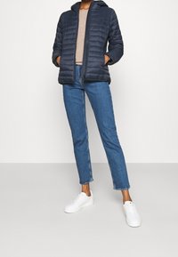 Navy puffer jacket with a hood, featuring quilted design and two side pockets. Paired with light denim jeans and white sneakers.