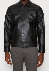 Homme portant une veste en cuir noire boutonnée avec des poches poitrines à rabat et un pantalon gris à motifs, se tenant devant un fond blanc.