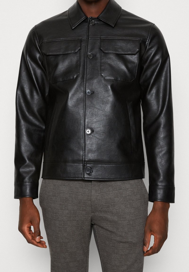 Homme portant une veste en cuir noire boutonnée avec des poches poitrines à rabat et un pantalon gris à motifs, se tenant devant un fond blanc.