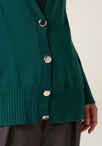 Cardigan teal con scollo a V, texture a coste e bottoni in tono argento. Presenta una vestibilità rilassata e maniche lunghe.