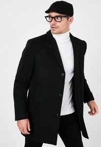 Black wool blend overcoat with a tailored fit, featuring a button closure, notched lapels, and two side pockets, worn over a white turtleneck.
