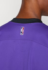 Ljubičasta košarkaška majica s crnim ovratnikom i NBA logotipom ušivenim na leđima. Tekstura tkanine s vidljivim detaljima šavova.