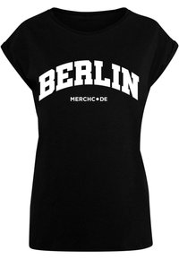 Musta t-paita, jossa on lyhyet käännetyt hihat, ja jossa on teksti "BERLIN" rohkeilla valkoisilla pääkirjaimilla ja "MERCH•DE" sen alla pienemmällä fontilla.