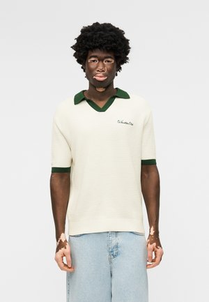 Νεαρός άνδρας με λεύκη που φοράει λευκό κοντομάνικο πλεκτό πόλο με πράσινο γιακά και ανοιχτό μπλε τζιν, όρθιος μπροστά σε απλό φόντο.