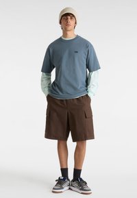 Camiseta azul de manga corta con logo negro, superpuesta sobre una camiseta de manga larga azul claro, combinada con pantalones cortos de cargo marrones y zapatillas grises.