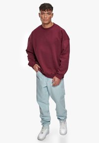 Weinroter Sweatshirt mit lockerer Passform und abgesenkten Schultern, kombiniert mit hellblauen Jeans und weißen Sneakers. Glattes Material und entspanntes Design.