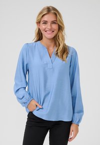 Blusa azzurro chiaro con scollatura a V e maniche lunghe, caratterizzata da polsini elasticizzati. Realizzata in un tessuto liscio con una leggera lucentezza.