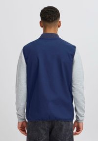 Muškarac okrenut leđima, obučen u mornarskoplavi bezrukavnik preko svijetlosivog dukserice s dugim rukavima i tamnosivih traperica, naslonjen na jednoličnu pozadinu.