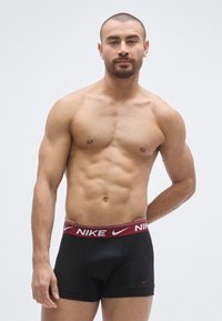 Μαύρα ανδρικά εσώρουχα Nike boxer με μπορντό ζώνη μέσης που φέρει λευκό λογότυπο. Το μοντέλο έχει μυώδη σωματότυπο και φαίνεται να στέκεται μπροστά σε ουδέτερο φόντο.