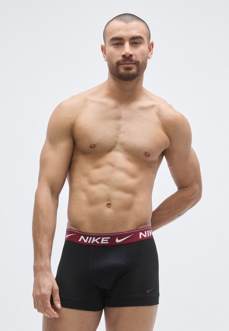 Μαύρα ανδρικά εσώρουχα Nike boxer με μπορντό ζώνη μέσης που φέρει λευκό λογότυπο. Το μοντ�έλο έχει μυώδη σωματότυπο και φαίνεται να στέκεται μπροστά σε ουδέτερο φόντο.