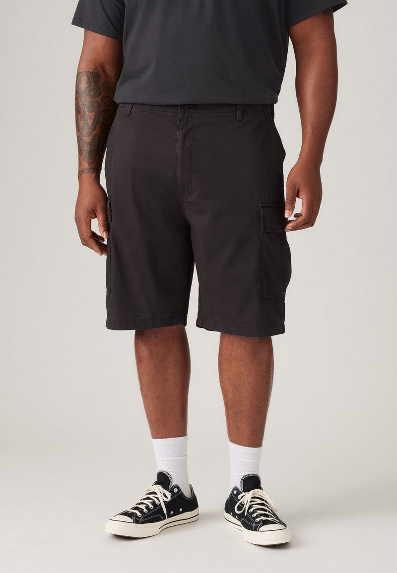  svarte cargo-shorts med sidelommer, slitesterkt stoff, rett ben-design, og en søm rett over kneet, parret med svarte joggesko og hvite sokker.