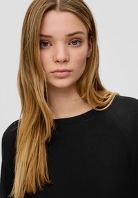Schwarzer Strickpullover mit Rundhalsausschnitt und Raglanärmeln, präsentiert von einer Person mit langen, glatten blonden Haaren und neutralem Make-up.