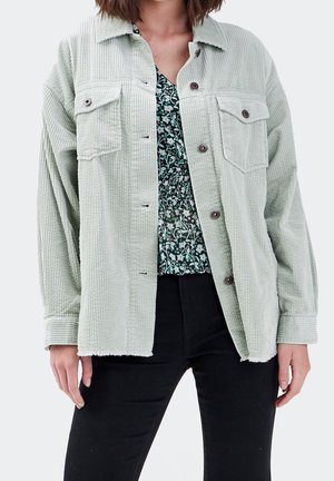 Femme portant une veste boutonnée en velours côtelé vert clair sur un haut à imprimé floral et un pantalon noir, debout sur fond blanc.
