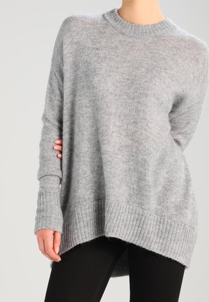 Femme portant un pull en tricot gris clair surdimensionné avec ourlet et poignets côtelés, associé à un pantalon noir, sur fond uni.
