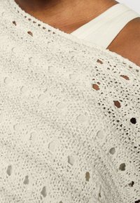 Gros plan sur une épaule à la peau brune portant un haut blanc en crochet par-dessus une bretelle de débardeur blanc.