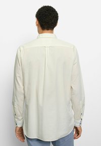 Hellbeiges Hemd mit Kragen und langen Ärmeln, weicher Textur, leichtem Glanz und blauen Knopfakzenten an Manschetten und Knopfleiste. Rückansicht dargestellt.