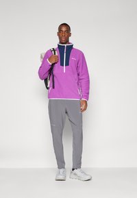 Lilla fleece pullover med marineblå accent, halv-lynlås foran og hvid kant. Sammen med grå træningsbukser og hvide sneakers.