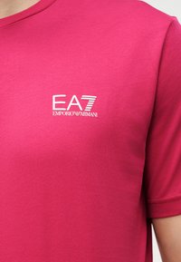 Φωτεινό ροζ πουκάμισο με κοντά μανίκια και λευκό λογότυπο EA7 Emporio Armani στο αριστερό μέρος του στήθους, που φοράει ένα άτομο.