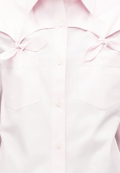 Chemise rose clair boutonnée avec deux poches poitrine, présentant des détails ajourés et des nœuds en tissu noués au-dessus de chaque poche.