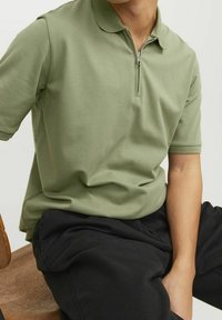 Polo tričko s krátkým rukávem ve světle zelené barvě, s límcem na zip a texturovanou tkaninou, doplněné černými kalhotami.