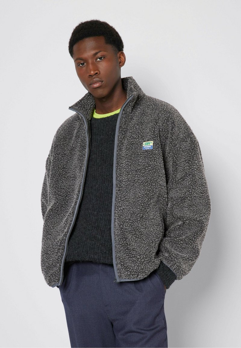 Grau Fleece-Jacke mit hohem Kragen, Reißverschluss vorne und einem grün-blauen Logo-Patch. Über einem dunklen Pullover und einer marineblauen Hose getragen.