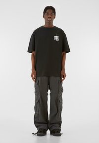 Sort T-shirt med hvidt logo, parret med løstsiddende grå cargo-bukser med flere lommer. Skoene er sorte med et lavprofil-design.