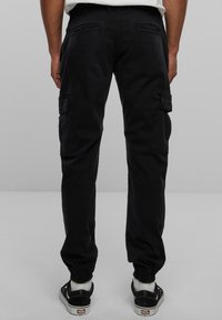 Pantalon cargo noir avec poignets élastiqués, comprenant deux poches latérales à rabat et des coutures contrastées. Tissu en coton texturé.