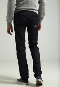 Jeans in denim scuro con vestibilità slim, dotati di una toppa a forma triangolare sulla tasca posteriore. Abbinati a una felpa grigia e scarpe nere.