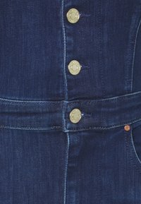 Mörkblå denimtyg med gyllene knappar och synlig sömnad. Har två övre knappar, en horisontell söm och en fickdetalj.