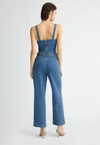 Džínový overall so širokými nohavicami, štruktúrovaným telom, nastaviteľnými popruhmi a pletenými detailmi v páse. Svetlomodrý denim s hladkou textúrou.