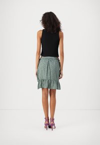 Schwarzes ärmelloses Oberteil, kombiniert mit einem grünen Rock mit Spitzenbesatz und Punkten, ergänzt durch lila Riemchen-Pumps.