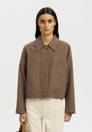 Mujer con cabello liso y oscuro lleva una chaqueta marrón con botones y pantalones crema, sosteniendo un bolso marrón claro sobre un fondo liso.