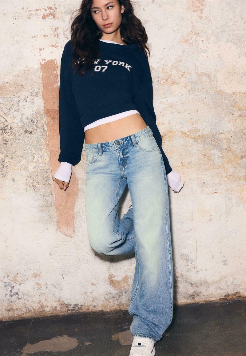 Giovane donna con capelli lunghi e scuri, che indossa una felpa corta blu navy e jeans a gamba larga azzurri, in piedi contro un muro testurizzato.