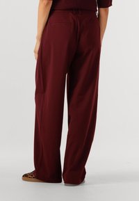 Bordeaux brede pijpenbroek met een gladde textuur, voorzien van achterzakken en een relaxte pasvorm. Opvallende schoenen met luipaardprint zichtbaar.