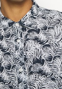 Camicia con colletto e bottoni, con motivo a foglia di palma blu navy e bianco, indossata da una persona con carnagione media.