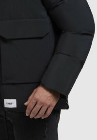 Zwarte pufferjack met een platte zak op de borst, elastische mouwen en een zichtbaar logo-label op de onderste zoom. Het materiaal lijkt glad en mat.
