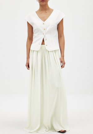 Biely crop top s krátkymi rukávmi a piatimi zlatými gombíkmi, v kombinácii s dlhou, voľne padajúcou svetlozelenou sukňou s plisovaním.