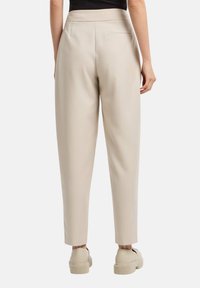 Pantaloni beige su misura con una texture liscia, vita alta e gamba affusolata; presentano una tasca posteriore e sono abbinati a scarpe neutre.