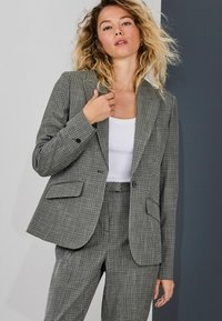 Hall triibuline bleiser, millel on lõhikuga disain, kaks eesmist taskut ja üks nupp. Kandes seda valge pluusi kohal, sobitatud samas triibulistes pükstega.