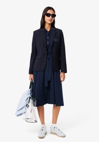 Blazer blu navy con un design aderente, indossato sopra un vestito blu navy abbinato. Gli accessori includono occhiali da sole neri e una borsa a righe. Sneakers bianche completano il look.