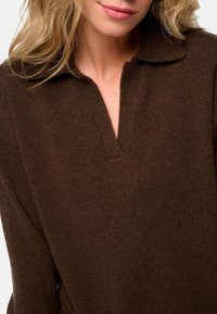 Pull en tricot brun avec un col et une finition texturée. Présente une fente à demi-boutonnage et des manches longues.