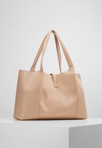 Beige texturerad läderväska med dubbla axelremmar och ett litet spänne, visad på en vit pelare.