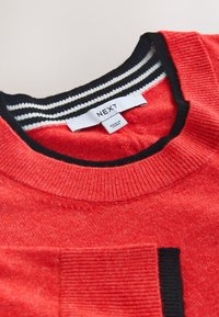 Pull en maille rouge avec col rayé noir et blanc et étiquette de marque « NEXT » cousue à l'intérieur de l'encolure, étiquette « Made in China » visible.