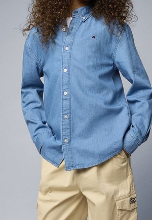 Person iført en lyseblå denimskjorte med knapper og hænderne i lommerne, kombineret med beige cargobukser med et lille logo-patch.